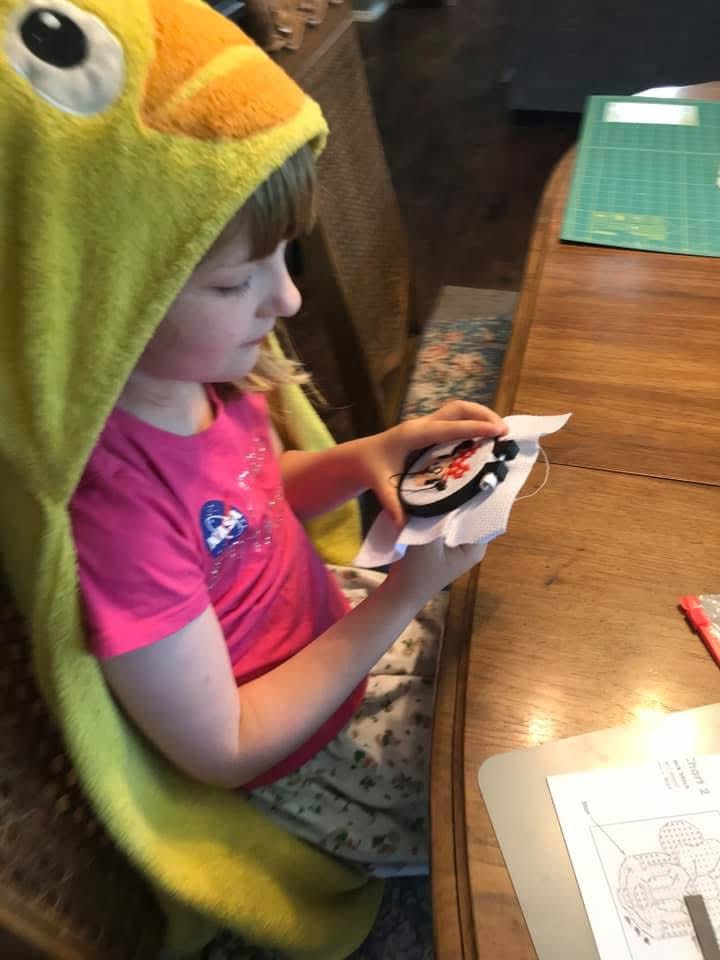 Makayla wearing a bright yellow duck hoodie while stitching a cross-stitch project at the dining table showing creativity at home.