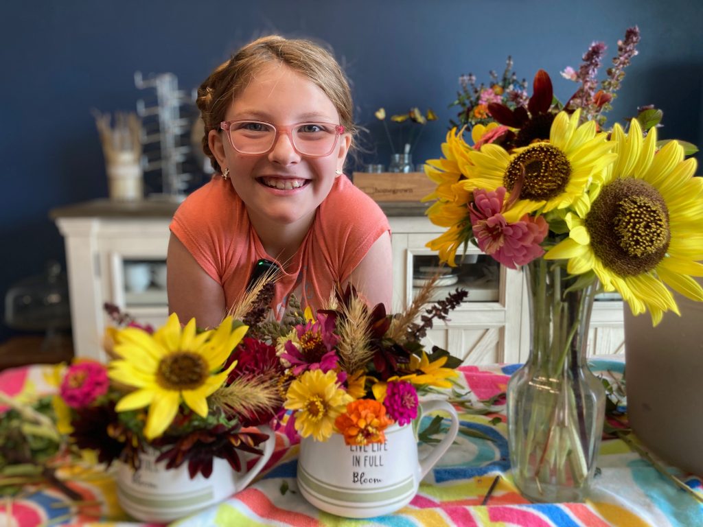 Makayla smiling behind colorful flower arrangements she created at home with sunflowers, zinnias, and grasses.